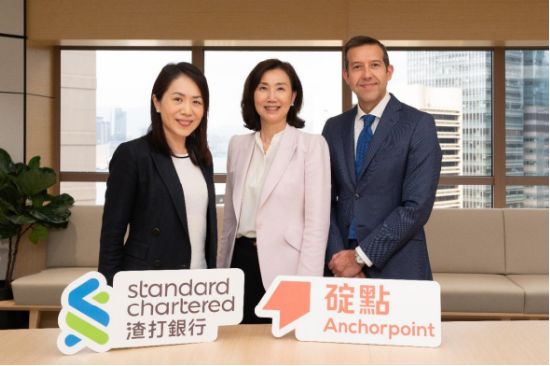 Three professionals—two women and one man in business attire—pose together in a modern office setting with a city skyline visible through the windows. In front of them are branded signs displaying "Standard Chartered 渣打銀行" and "碇點 Anchorpoint," visually representing the partnership between Standard Chartered and Anchorpoint related to the HKDAP stablecoin initiative mentioned in the post.