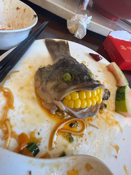A partially eaten fish head with corn on the cob stuffed in its mouth sits on a dirty plate alongside watermelon rind and chopsticks.