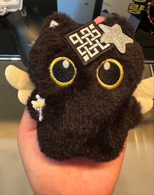 A hand holds a small, round, black plush toy with big yellow eyes, a star patch, and a decorative patch with a maze-like symbol.