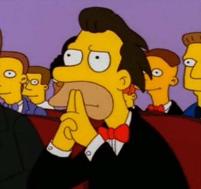 An animated scene from The Simpsons shows a mustachioed man in a tuxedo making a 'shh' gesture with his finger over his mouth while sitting in a crowd.