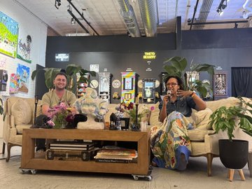 Two people sit in armchairs on a stage in what appears to be a podcast studio, with framed pictures on the wall behind them and plants decorating the space.