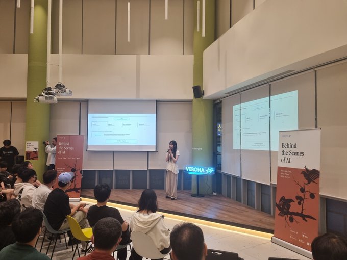 A woman speaks on stage at a Verona crypto event titled "Behind the Scenes of AI" in Seoul, with a presentation slide and branded banners visible behind her as the audience watches attentively. The setting reflects the @verona_dev talk referenced in the post, capturing the moment just before the $VERONA token airdrop that drew cheers from attendees at KBW2025.