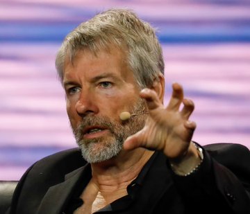A close-up photo of Michael Saylor, the MicroStrategy chairman known for Bitcoin advocacy, speaking with a headset microphone while gesturing with his hand.