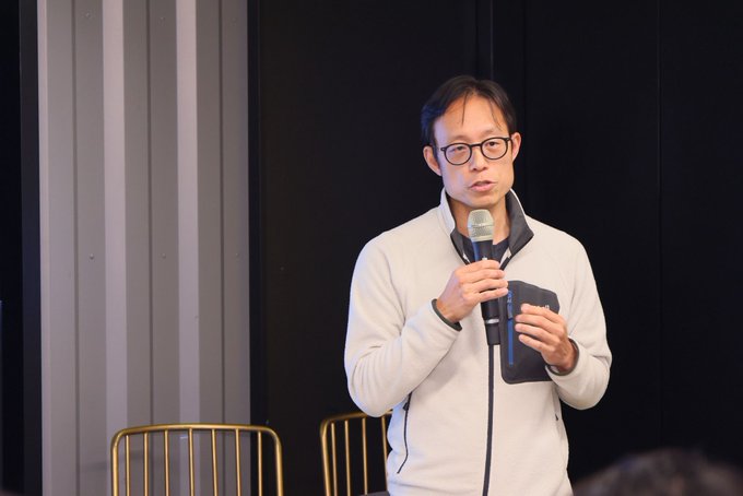 A man wearing glasses and a cream-colored zip-up sweater holding a microphone and a notebook, appearing to speak at an event or presentation.