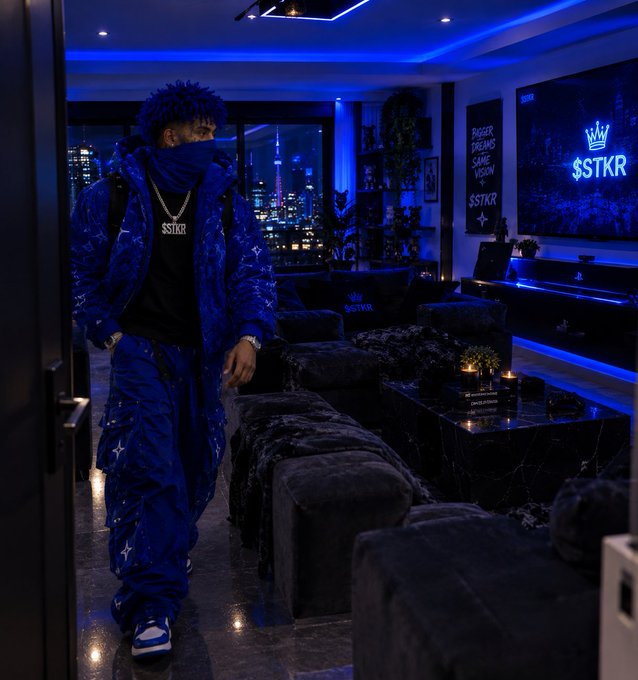 A person with dreadlocks wearing a blue patterned tracksuit and a '$TKR' necklace stands in a modern, blue-lit room with a '$TKR' neon sign and framed wall art.