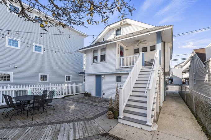 A real estate photo shows the back of a white two-story house with an elevated deck accessed by a staircase, a stone patio with a dining set, and string lights overhead.
