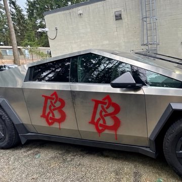 A silver Tesla Cybertruck parked outside with red graffiti that reads '18 18' sprayed on the door.