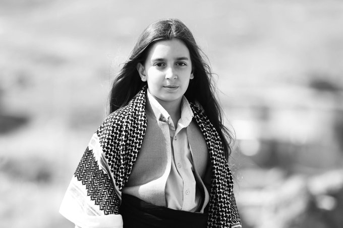A black-and-white portrait of a young woman with long hair wearing a houndstooth vest over a button-up shirt, with the name 'kazuhi' credited.