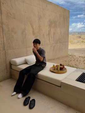 A man in a gray shirt and dark pants sits on a beige outdoor sofa in a minimalist concrete room with an open wall revealing a desert landscape, with a tray of food and slippers placed nearby.