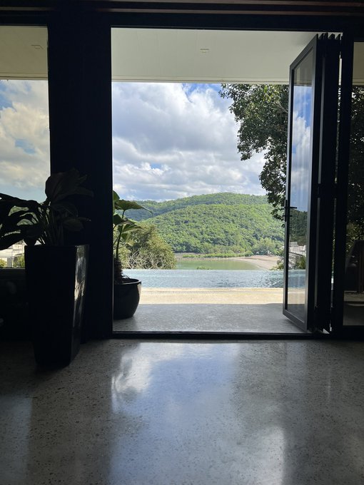 A view from inside a modern home looking out through open glass doors toward a green mountain and body of water, with a potted plant on the polished floor.