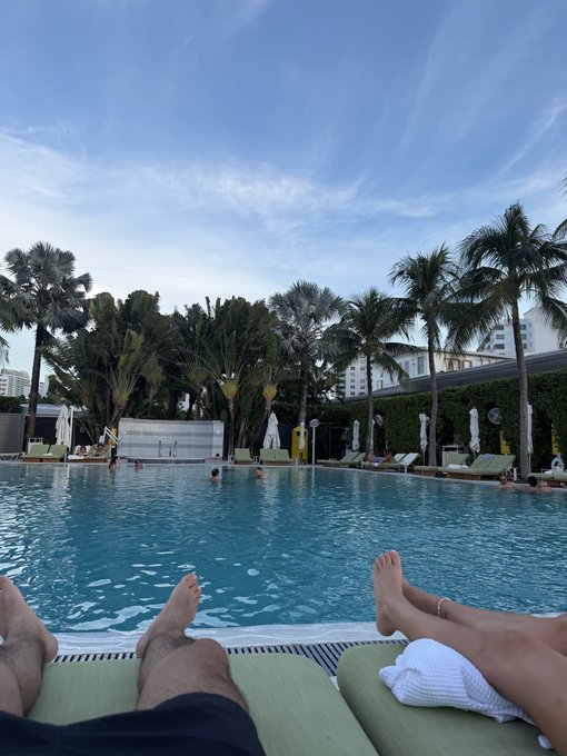 A couple relaxes by a tropical resort pool, feet propped up on green lounge chairs as palm trees sway against a serene blue sky. The scene perfectly captures a steady, laid-back vacation vibe—matching the "Steadily vibin" mood with its tranquil poolside atmosphere.