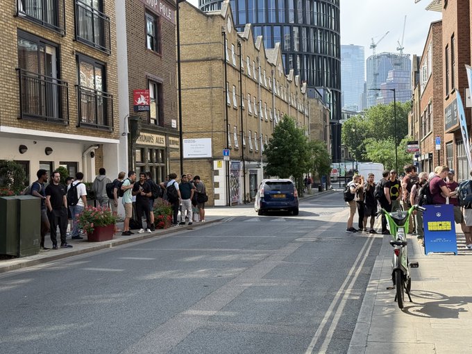 A crowd gathers outside an event venue for Arbitrum's Open House, with attendees lined up along a London street under sunny skies. Visible signage includes a "YOUNG'S" pub sign and a motorcycle parking sign, setting the scene of a bustling tech and business-focused gathering.
