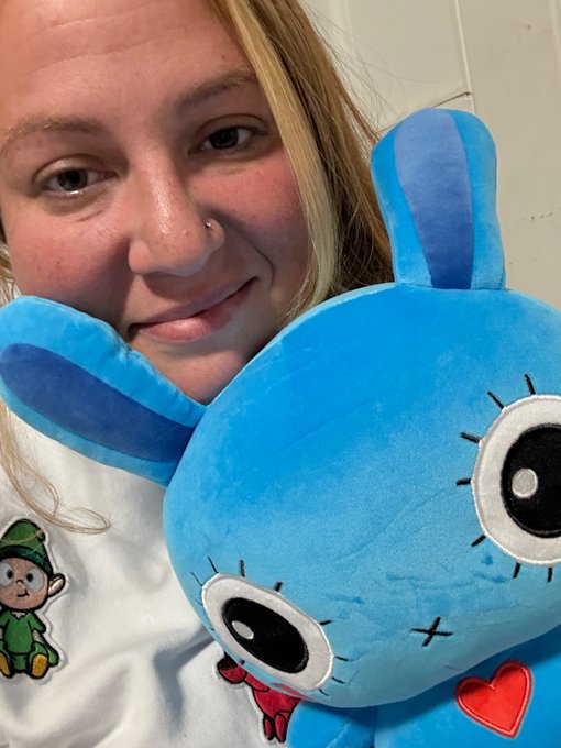 A young woman with blonde hair and a nose piercing smiles while holding a large blue plush bunny toy with big eyes and stitched details.