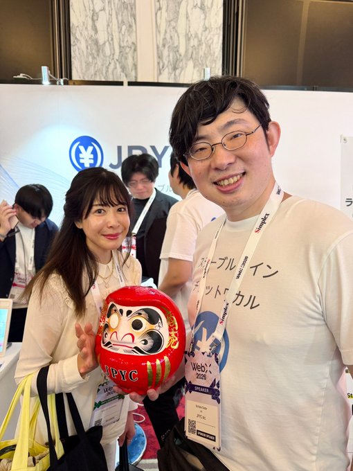 Two smiling attendees at #WebX 2025 pose with a red Daruma doll displaying "JPYC" branding, promoting the Japanese yen stablecoin at a Stripe booth, illustrating the post's claim that $JPYC can now be stored in a hardware wallet. The man wears a conference lanyard and a shirt with Japanese text including "Japanese" and "Stablecoin," tying directly to the announcement of secure self-custody options for the yen-pegged token.