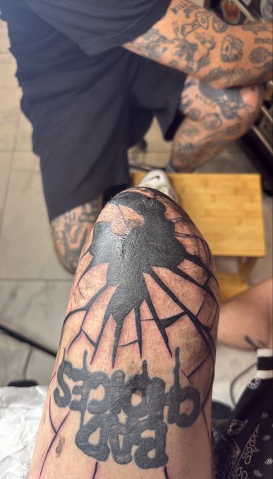 A person's leg with a large black tattoo of a cross and stylized lettering, freshly inked, with another heavily tattooed person sitting in the background.