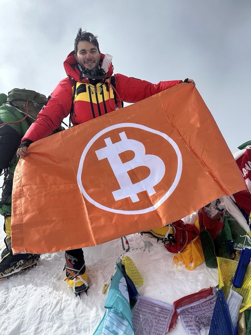 A climber in heavy cold-weather clothing proudly displays a bright orange flag featuring the Bitcoin (₿) logo while standing atop a snow-covered summit surrounded by colorful prayer flags. The image brings the accompanying caption "Bitcoin flag on top of Mount Everest" to life, capturing the symbolic milestone of cryptocurrency reaching the world's highest peak.