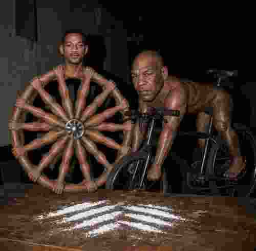 The image shows a bizarre, darkly humorous scene featuring two shirtless men on a dimly lit stage—one positioned inside a large spinning wheel-like structure made of multiple human arms radiating outward like spokes, while the other rides a bicycle toward him, suggesting an absurd performance or stunt. In the foreground, a wooden surface displays white powdery markings resembling a racetrack or playing field, reinforcing the caption "Choose your player" as the men appear to be contestants in a surreal, high-stakes game. The overall mood is gritty and unsettling, blending athleticism with grotesque spectacle.