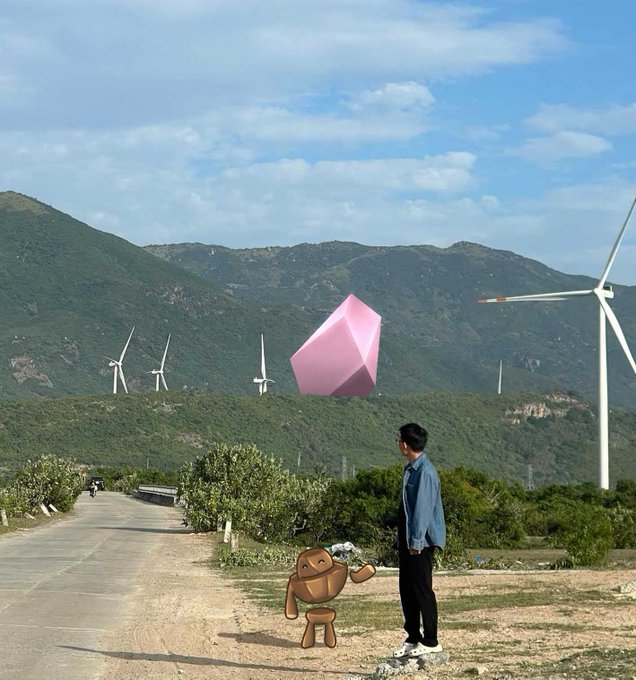 A person in a blue shirt stands on a path with a small brown robot-like figure, looking toward a massive floating pink geometric object hovering in the sky against a mountainous landscape with wind turbines.