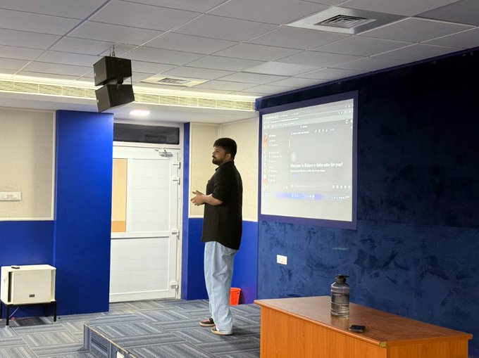 A man stands in a blue-walled lecture hall presenting next to a projected slide that reads 'Welcome to the Machine Learning for the Rest of Us' workshop.