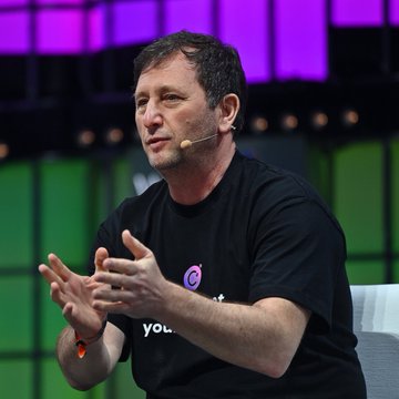 A man with short dark hair, wearing a black t-shirt with a purple logo and the partial text "you." visible, speaks into a headset microphone while gesturing with his hands on a stage with purple and green lighting. The image accompanies news about federal prosecutors opposing former Celsius CEO Alex Mashinsky's attempt to vacate his 12-year prison sentence.