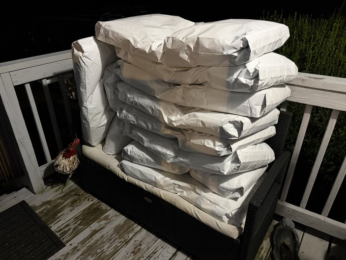A stack of multiple white cushions/pillows piled on top of each other on a wicker sofa on an outdoor porch or deck at night, with a decorative rooster figurine visible to the left.