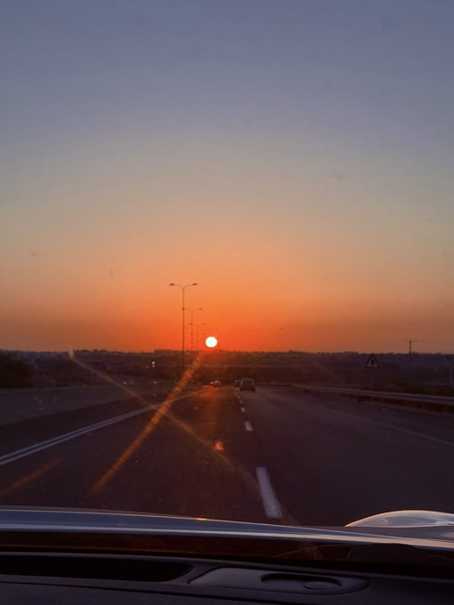 A view from inside a car at sunset shows the sun setting low on the horizon over a highway, with other vehicles visible in the distance.