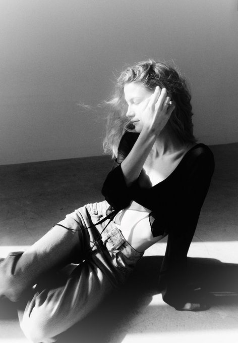 A black and white photo of a woman sitting on the floor, eyes closed, with her hand gently touching her face, wearing a dark top and light pants.