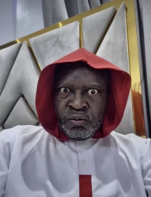 A person with a serious expression wearing a white robe and a red hood, with what appears to be a small cross on their chest.