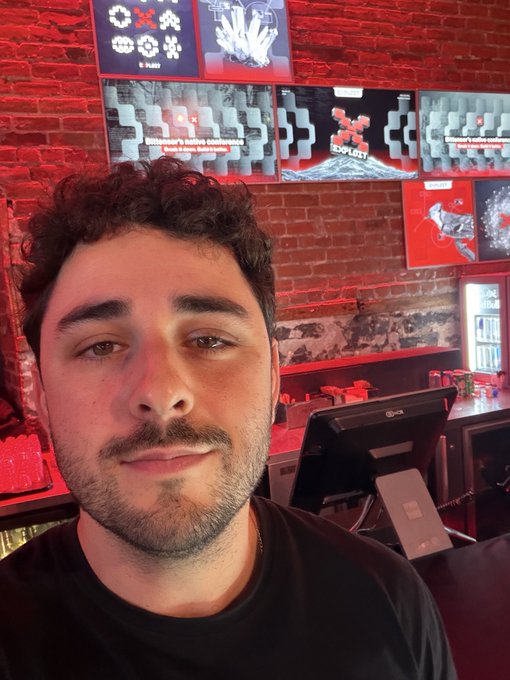 A selfie of a young man with dark curly hair and a short beard, taken inside a restaurant with red ambient lighting and menu boards displaying food items on the brick wall behind him.