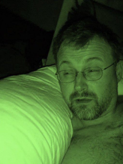 A nighttime selfie of a man with glasses and a beard, with a greenish tint, resting his head against a white pillow or blanket.