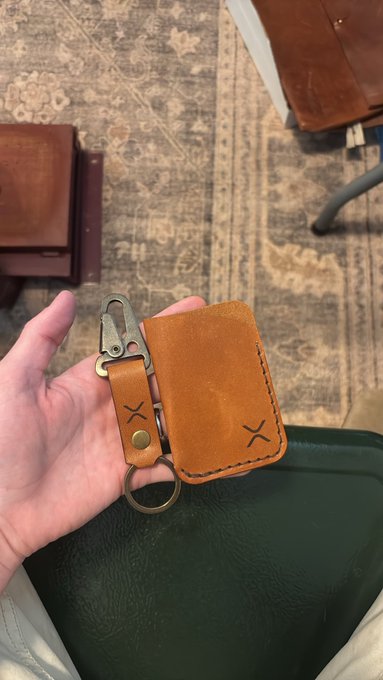 A hand holding a tan leather key organizer with a brass key clip and ring, featuring an 'X' logo, photographed against a patterned rug.