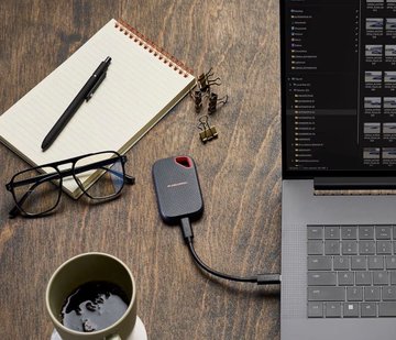 A small gray Sandisk portable SSD rests on a wooden desk next to a laptop displaying lines of code, a spiral notebook with a pen, eyeglasses, and a cup of coffee—visually representing the device claimed to hold 70 years of Bitcoin blockchain data, despite ongoing debates about running nodes.