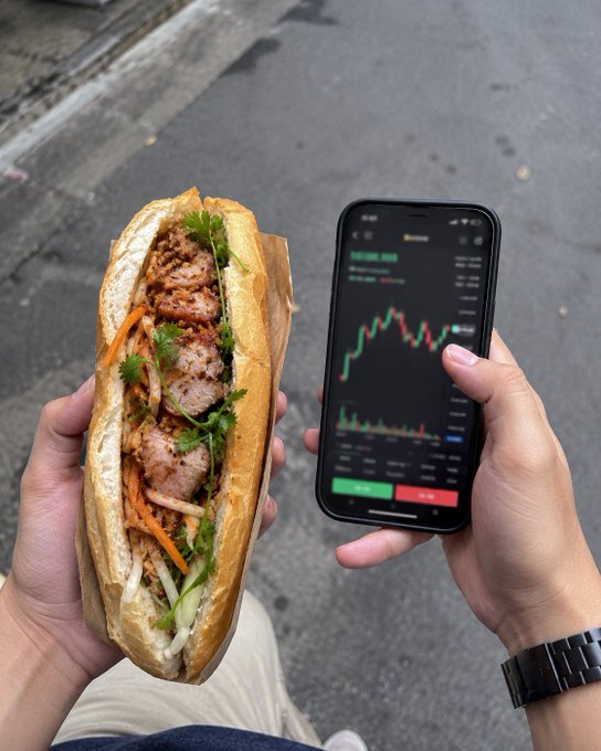 A first-person view of hands holding a Vietnamese banh mi sandwich in one hand and a smartphone displaying a green stock or crypto chart in the other.