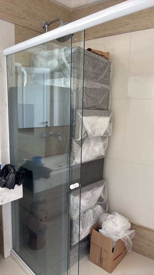 A photograph of a bathroom under construction or renovation, featuring a glass shower enclosure, a bathtub, hanging grey storage organizers, and bubble wrap scattered on the floor.