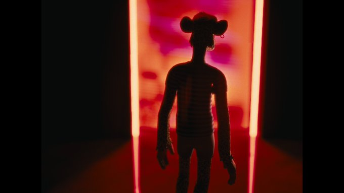 A stylized silhouette of a person with mouse-like ears standing in a doorway illuminated by dramatic red lighting, with tattoos visible on their arms and hands.