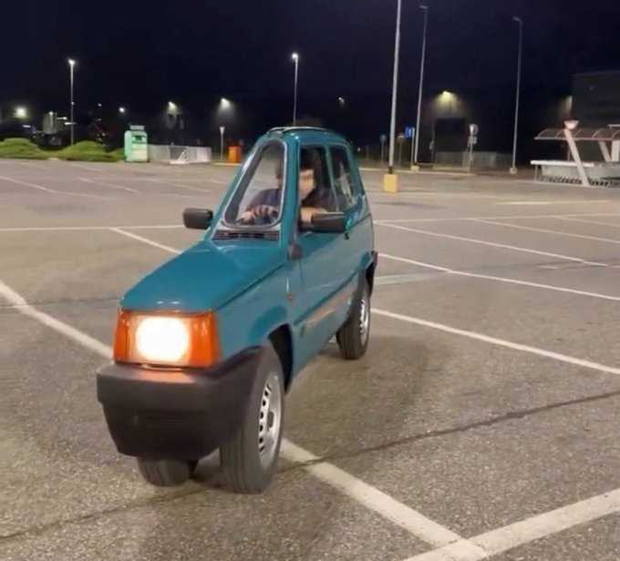 A person driving a very small teal compact car through an empty parking lot at night, with streetlights visible in the background.