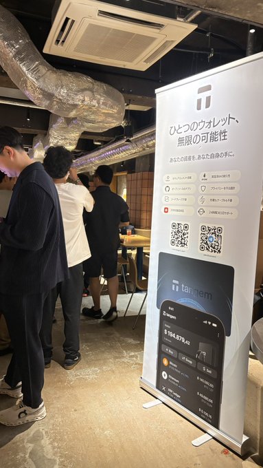 The image shows a Tangem-branded promotional banner displayed at what appears to be a crypto event, with attendees gathered nearby. The banner highlights Tangem's wallet features in Japanese and displays a phone screen showing crypto balances including "1166.8770 XRP," connecting to the post about the Flare × Tangem × XRP event.