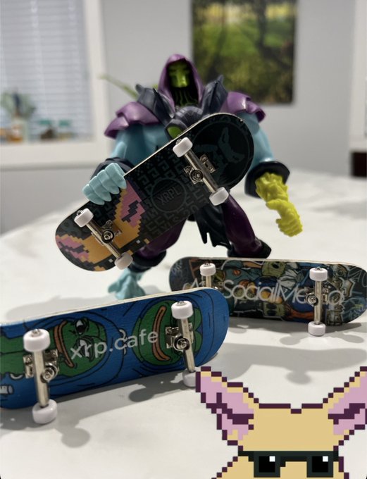 A Skeletor action figure stands behind three fingerboards (mini skateboards) on a white surface, with a small pixel art character in the bottom right corner.