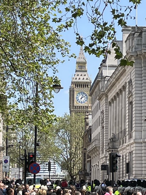 Big Ben clock tower is visible through spring foliage along a busy London street, with crowds of pedestrians gathered below.