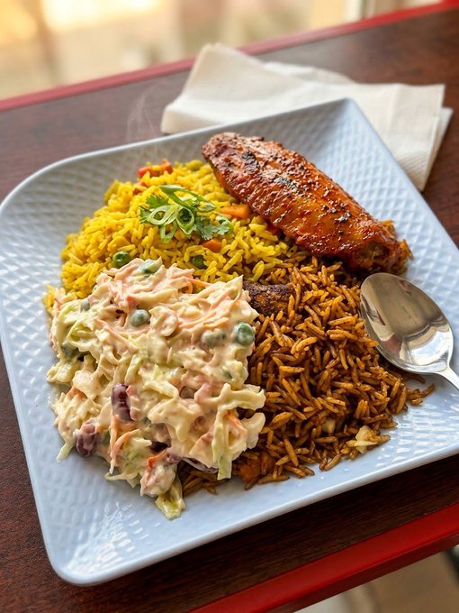 A white plate holds a trio of dishes—yellow rice topped with a glazed chicken sausage, a dark seasoned rice, and creamy coleslaw—alongside a spoon, though the caption notes the protein source was unaffordable eggs.