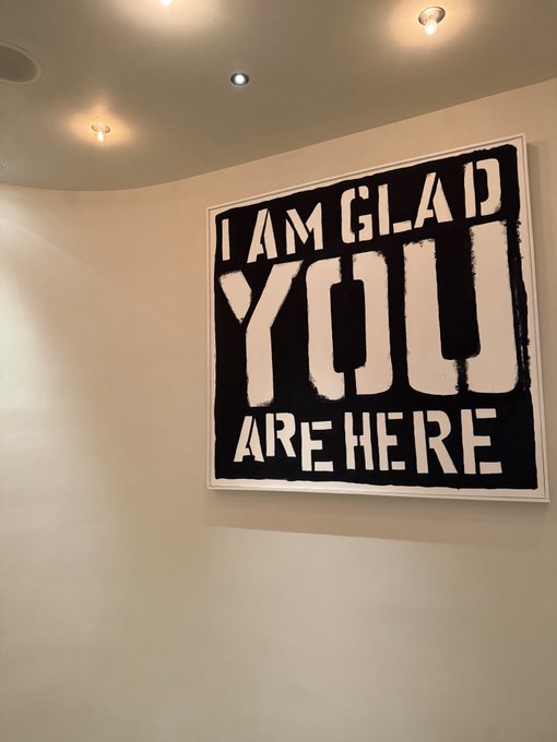 A framed black-and-white artwork mounted on a white gallery wall displays the hand-painted text 'I AM GLAD YOU ARE HERE'.