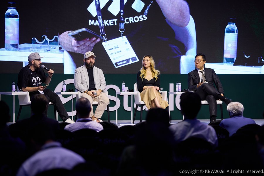 A panel discussion takes place at a "KBW2026" event with four speakers seated on stage in front of a large screen displaying "MARKET CROSS," with audience members visible in the foreground.