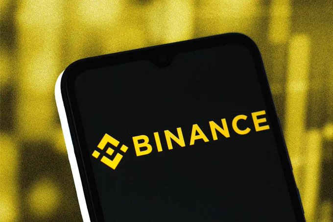 A smartphone displays the yellow "BINANCE" logo against a dark screen, with a blurred yellow Binance-branded background. The image relates to the post by visually representing Binance, the cryptocurrency exchange announcing its update to commodity-based TradFi perpetual contracts switching to a 24/5 trading model.