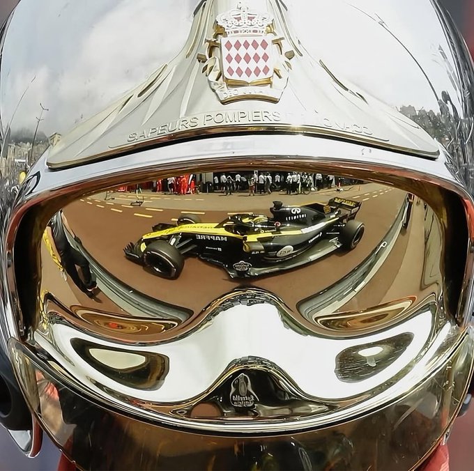 A fisheye reflection on a chrome Sapeurs Pompiers (French firefighters) helmet shows a Renault Formula 1 race car, with the Monaco coat of arms visible on the front.