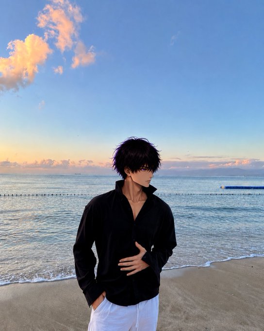 An anime-style illustration depicts a dark-haired young man in a black shirt and white pants standing on a beach during sunset.