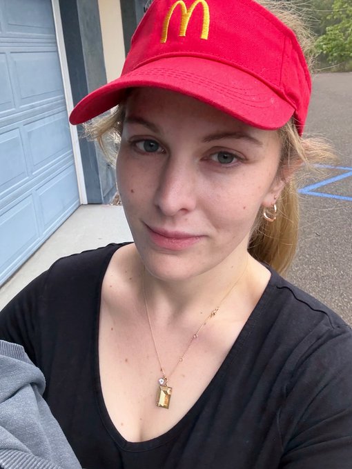 A young woman with blonde hair takes a selfie wearing a red McDonald's cap with the golden arches logo, a black v-neck top, and a delicate necklace with a gold pendant, standing outside a garage.