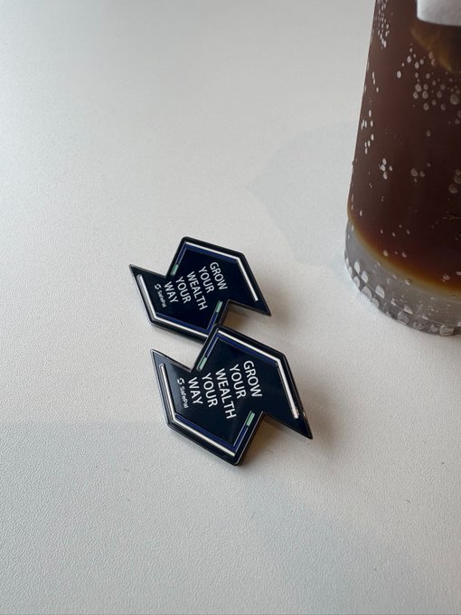 Two arrow-shaped enamel pins with the inspirational text "GROW YOUR WEALTH YOUR WAY" lie on a white surface beside a dark beverage, symbolizing a new week of boldly pursuing opportunities and growing wealth on one's own terms.