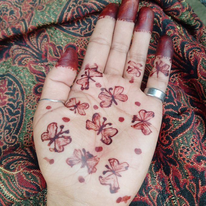 A close-up of a hand with intricate dark red henna designs featuring butterfly and floral motifs, along with red dye staining the fingertips, resting against a richly patterned red, green, and black fabric.