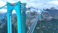 A vibrant turquoise suspension bridge spans a misty mountain valley, resembling the iconic architecture of China's Guizhou region. The image humorously contrasts with the caption's playful suggestion that the university has expanded to China, using the striking, exotic landscape to imply a far-flung academic outpost.