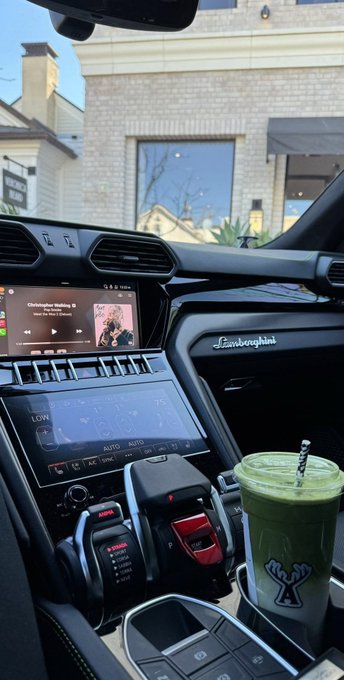 Interior view of a Lamborghini car dashboard showing the infotainment screen, air controls, and a green iced matcha drink in a cup holder, with a house visible through the windshield.